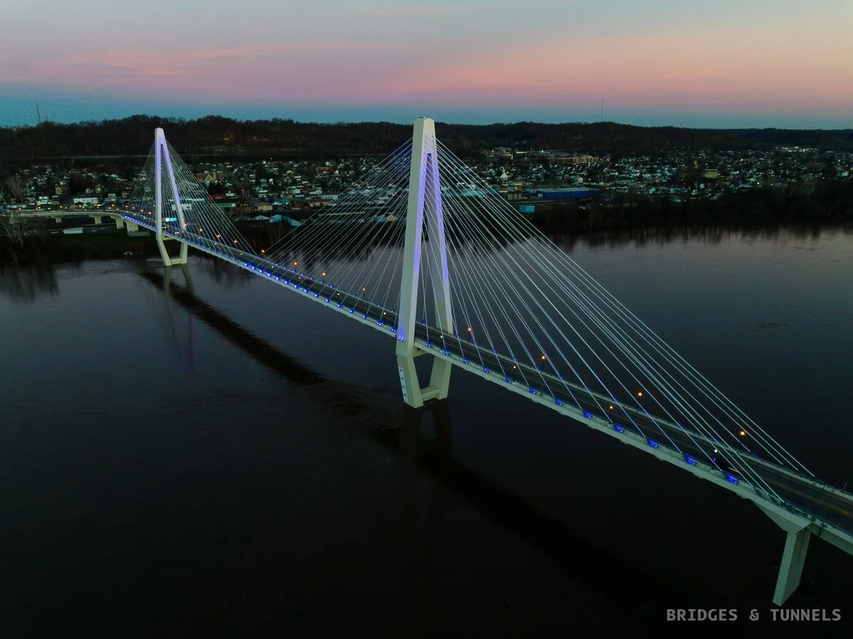 An illuminated cable-stayed bridge over the Ohio River at dusk, with Ironton, OH skyline in the background.