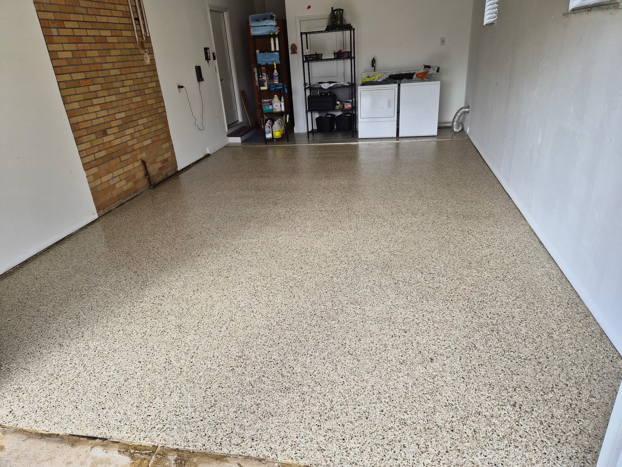 Empty garage with speckled epoxy floor, brick wall, white walls, laundry area with washer and dryer, black shelving unit, and a brick wall section with hooks.