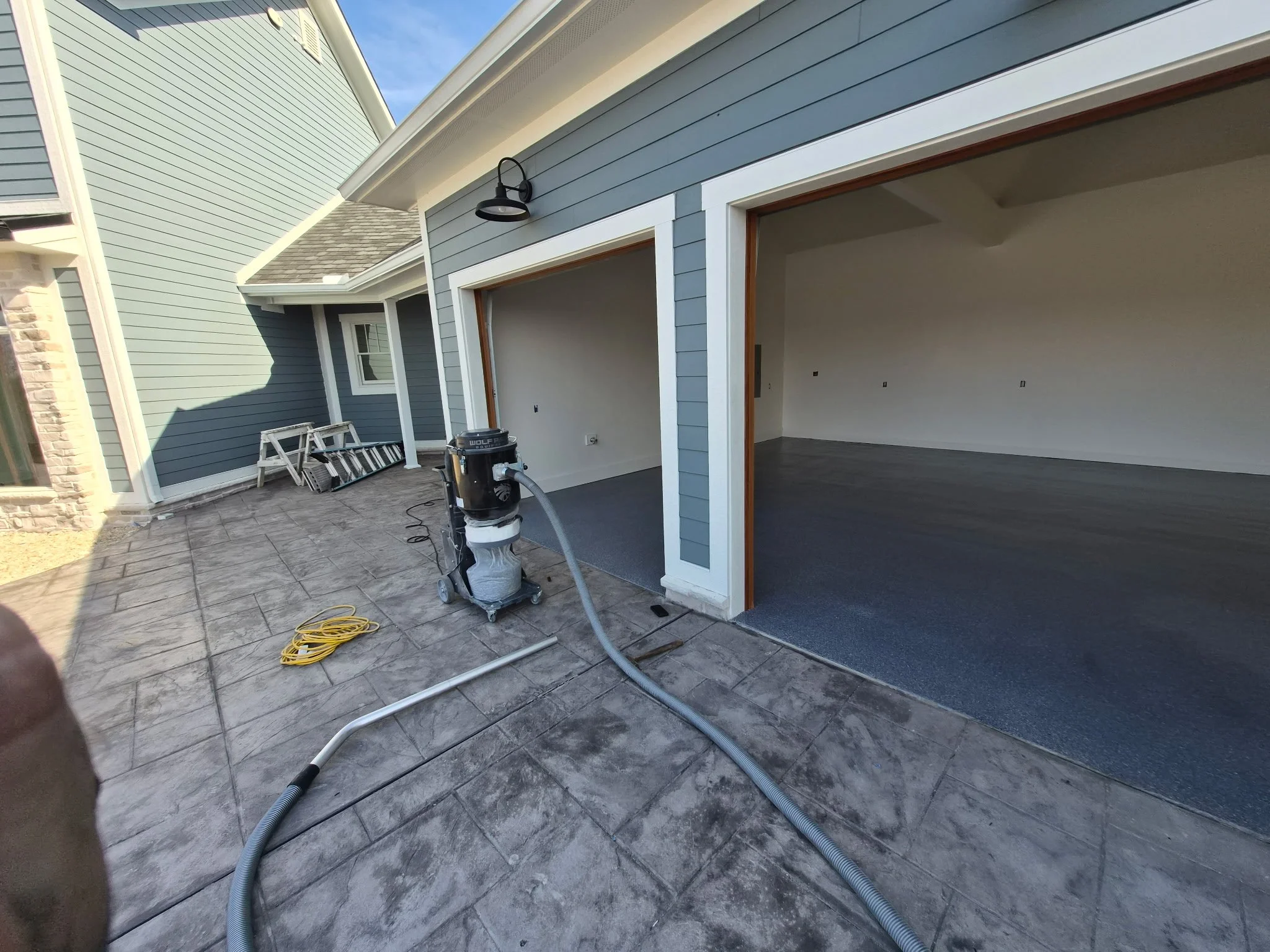 View of an outdoor patio area with stamped concrete flooring and an unfinished garage with open doors, showing interior walls and flooring, with some construction equipment and ladder leaning against the house.
