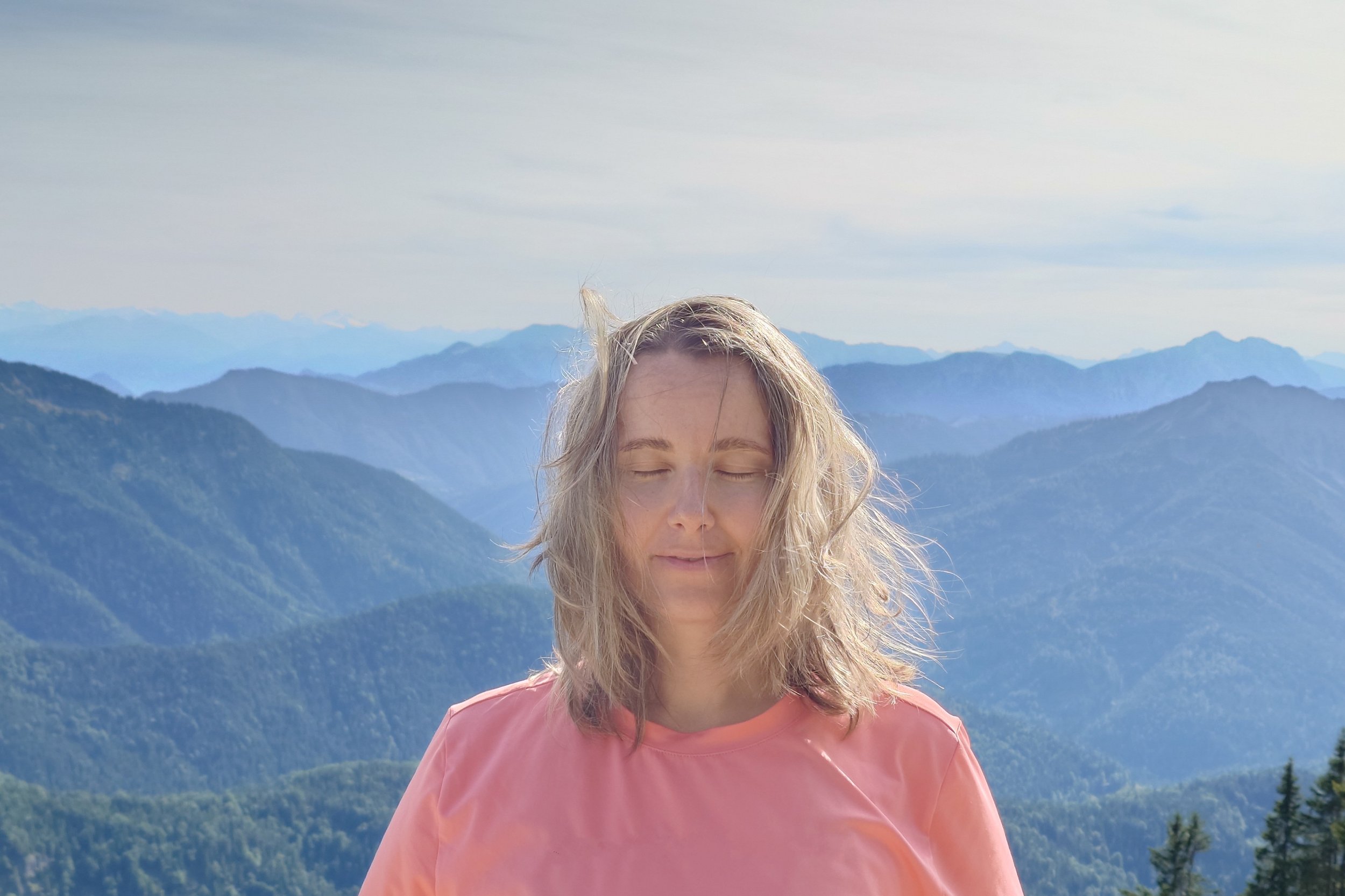 Woman with blonde hair in a pink shirt enjoying the mountain view with her eyes closed.