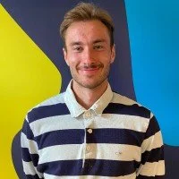 A man with light brown hair and a beard smiling, wearing a blue and white striped sweater, standing in front of a colorful background with yellow, blue, and black shapes.