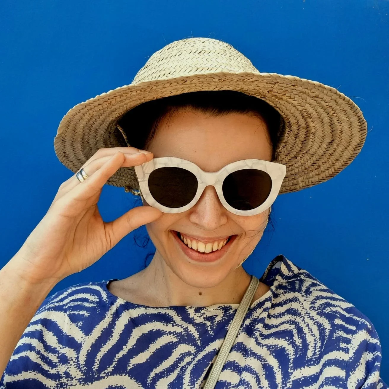 A woman wearing a straw sun hat, white sunglasses, and a blue and white patterned top smiling against a blue background.