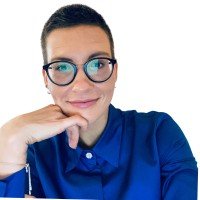 A woman with short dark hair, glasses, and a blue shirt resting her chin on her hand, smiling at the camera.