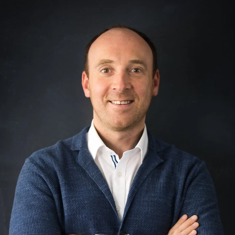 A portrait photograph of a smiling man with short brown hair, wearing a white collared shirt and navy blue sweater, standing against a dark background.