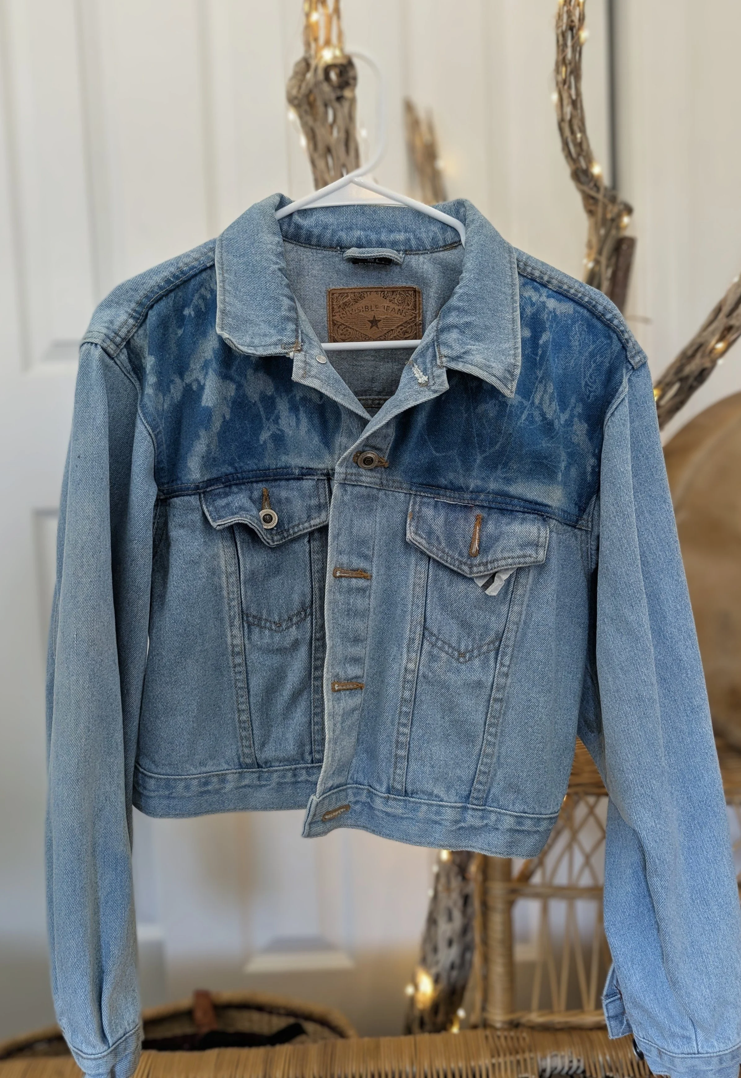 A blue denim jacket hanging on a white hanger, with some distressing details, in front of a decorative wall with branches and lights.