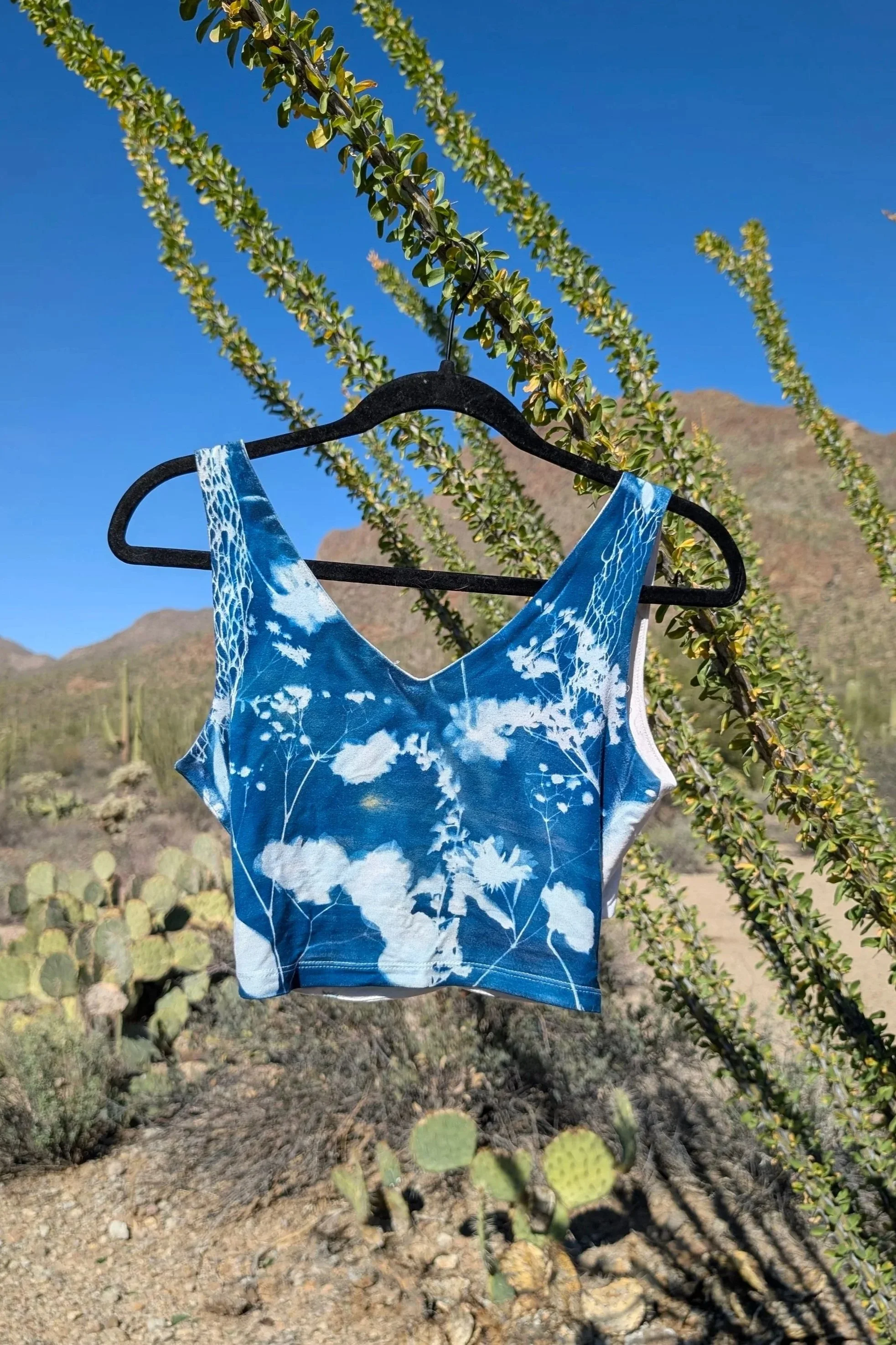 A blue tie-dye crop top with white floral pattern hanging on a black hanger outdoors against a desert background with cacti, bushes, and mountains under a clear blue sky.