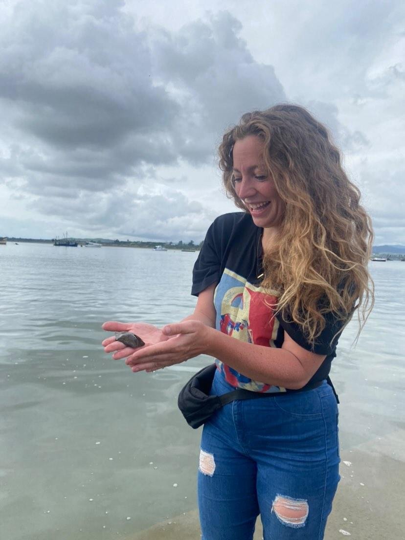 A woman with curly hair standing in waist-high water at the beach, holding a small turtle in her hands and smiling happily under a cloudy sky.