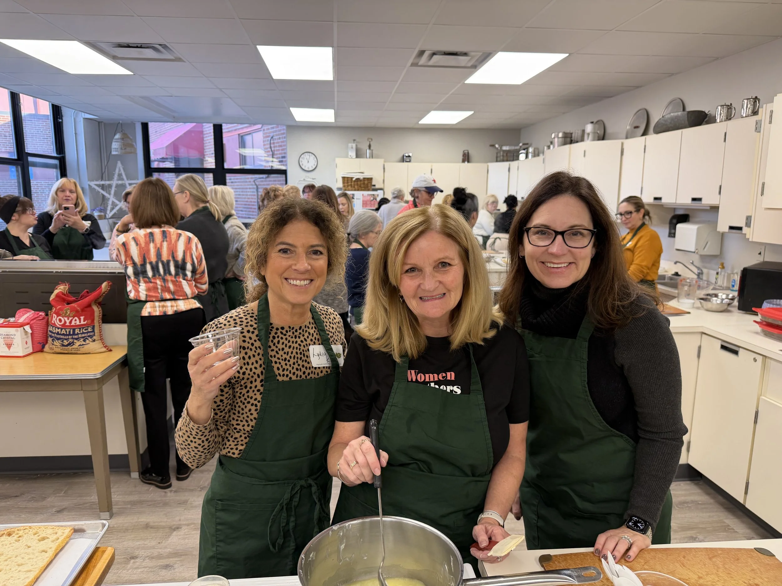 Three women stand together in a kitchen, smiling at the camera. They are wearing green aprons and preparing food. The background shows a busy room with many people, kitchen cabinets, and a large window.