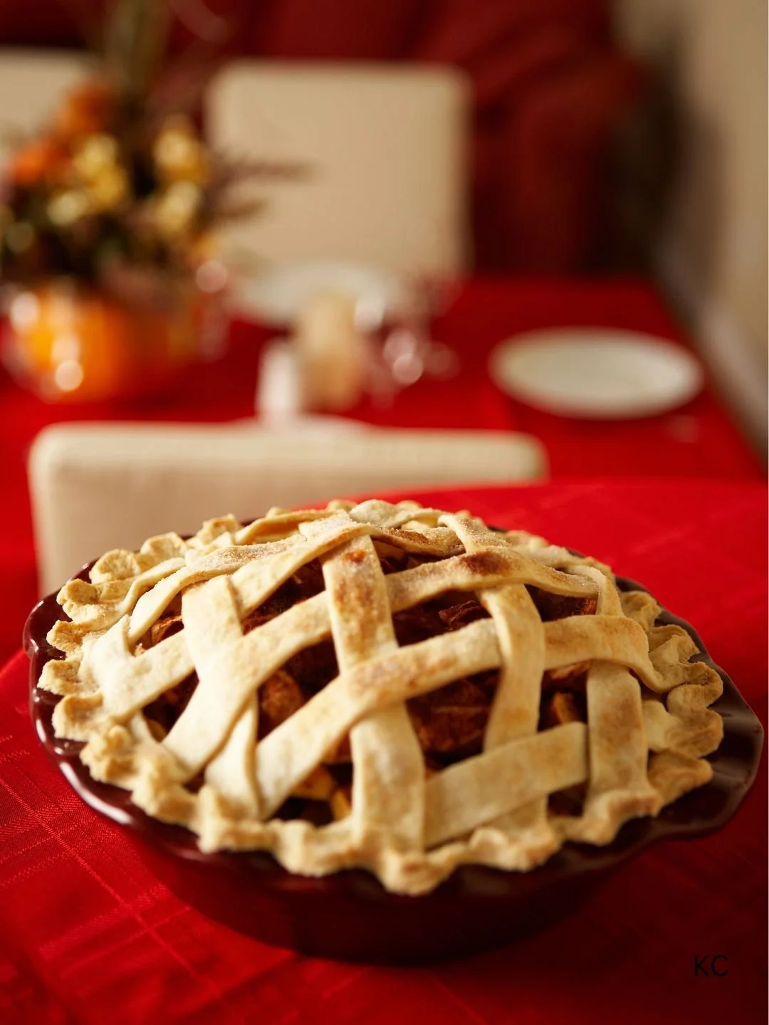 Pie with a lattice crust on a dark plate on a red tablecloth, with a blurred holiday table setting in the background.