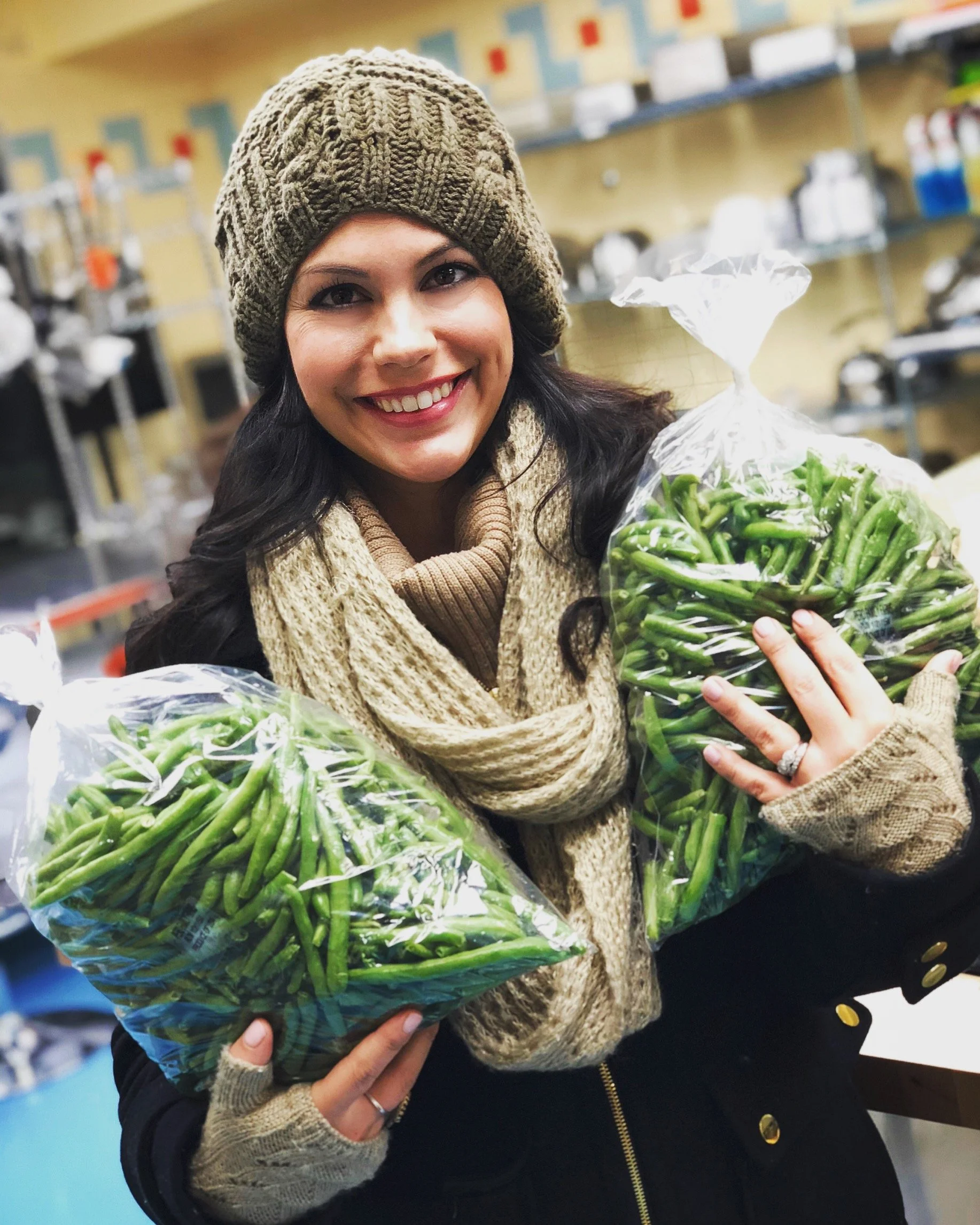 A smiling woman in warm clothing holding two bags of green beans in a grocery store.