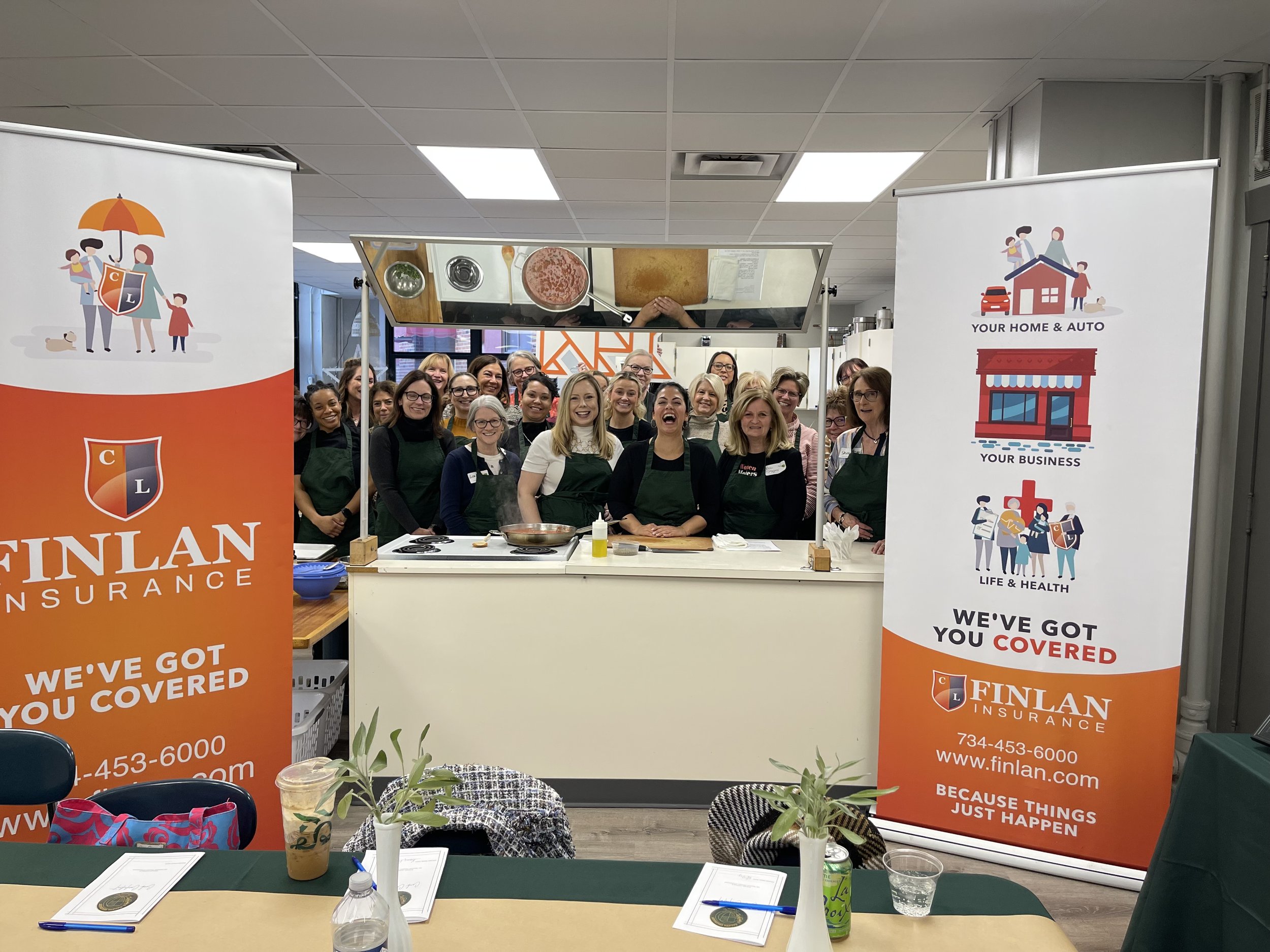 Group of people wearing aprons gathered behind a kitchen counter in a cooking class or event, smiling at the camera. Two large banners with the Finlan Insurance logo and services stand on either side of the group. The setting appears to be a commercial kitchen or cooking studio.