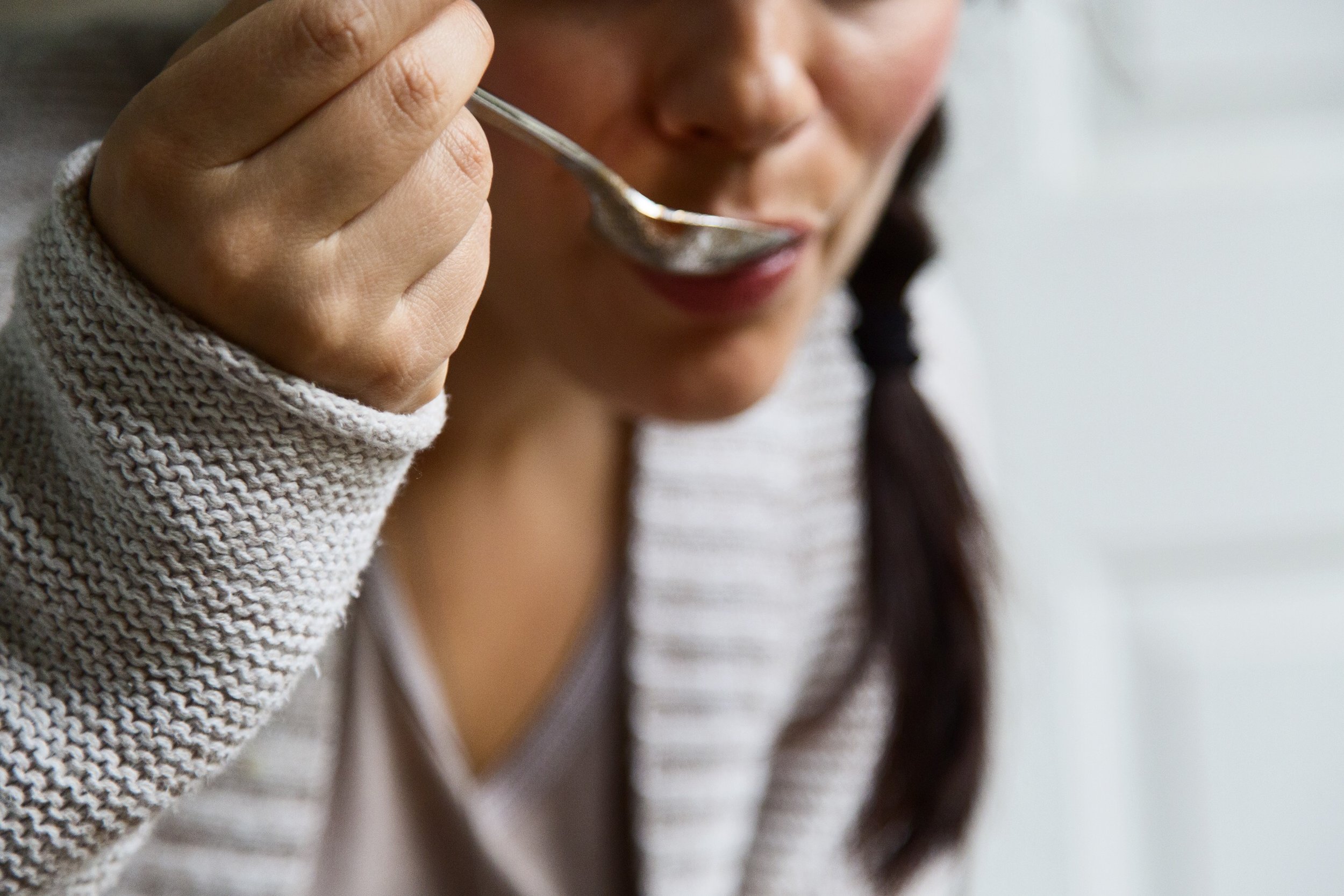 Person wearing a beige knitted sweater, holding a spoon near her mouth, about to taste food.