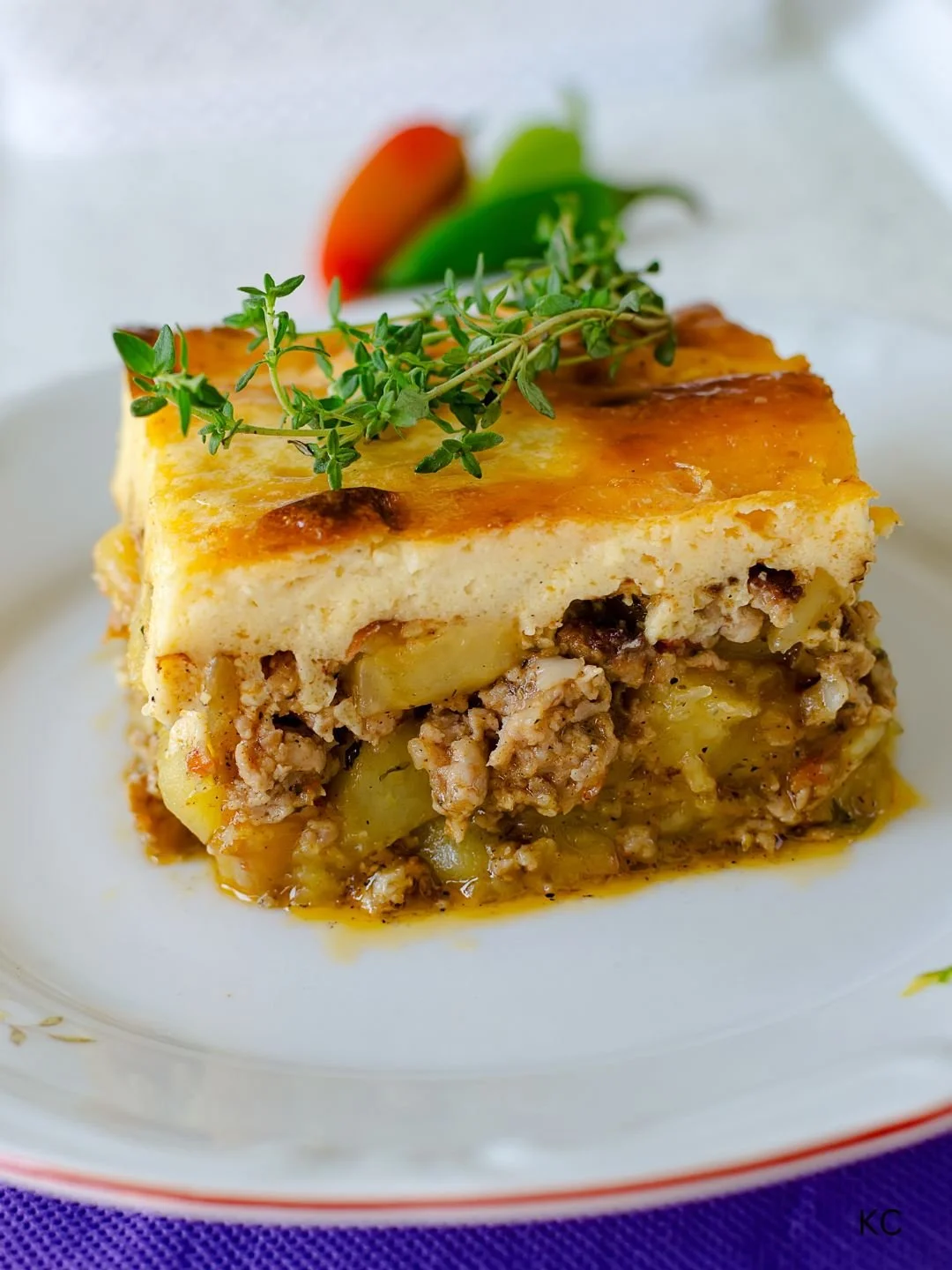A slice of casserole with layers of ground meat, potatoes, and cheese, garnished with fresh thyme and served with sliced tomatoes and peppers in the background.