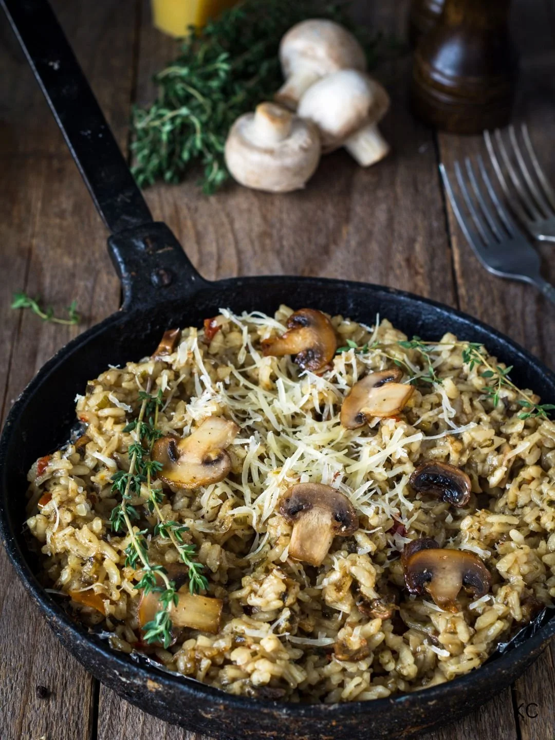 Cooked mushroom risotto garnished with cheese and herbs in a skillet, with fresh mushrooms and herbs on a wooden table.