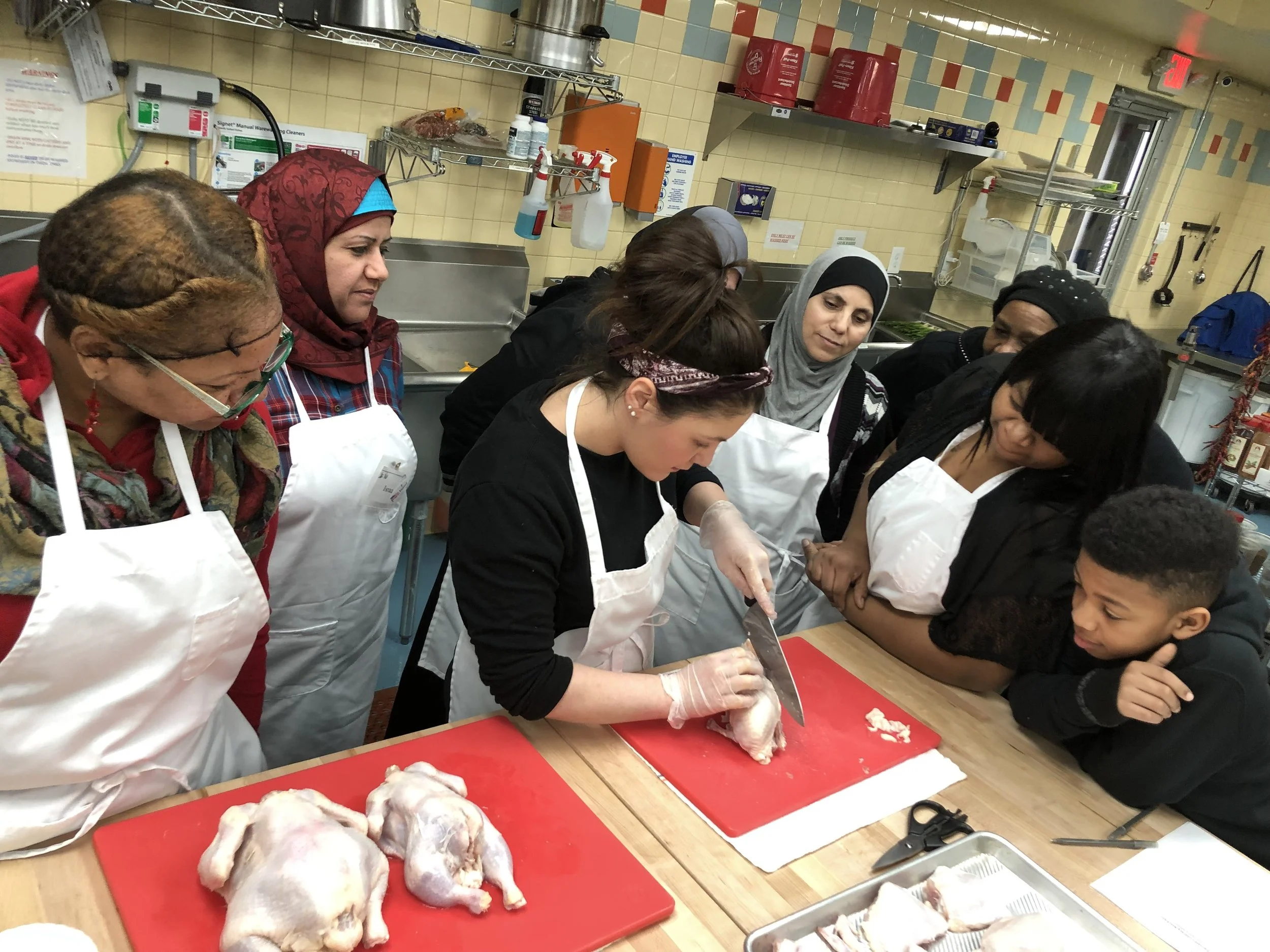 A group of women and a boy gathered around a kitchen counter watching a woman cut a raw chicken with a large knife, with chickens on a red cutting board in the foreground.