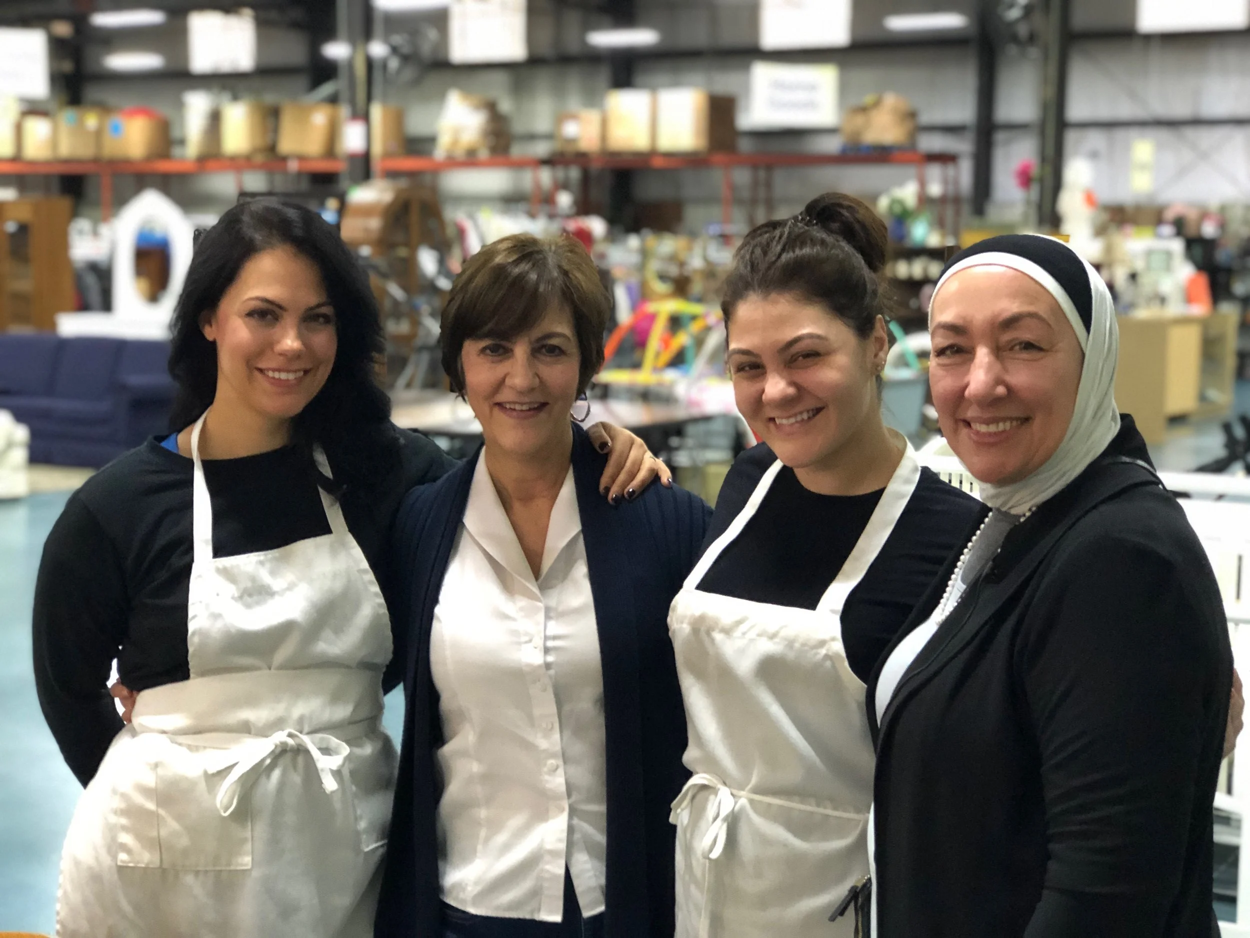 Four women standing together in a warehouse or store, smiling at the camera, some wearing aprons, surrounded by shelves of various items.