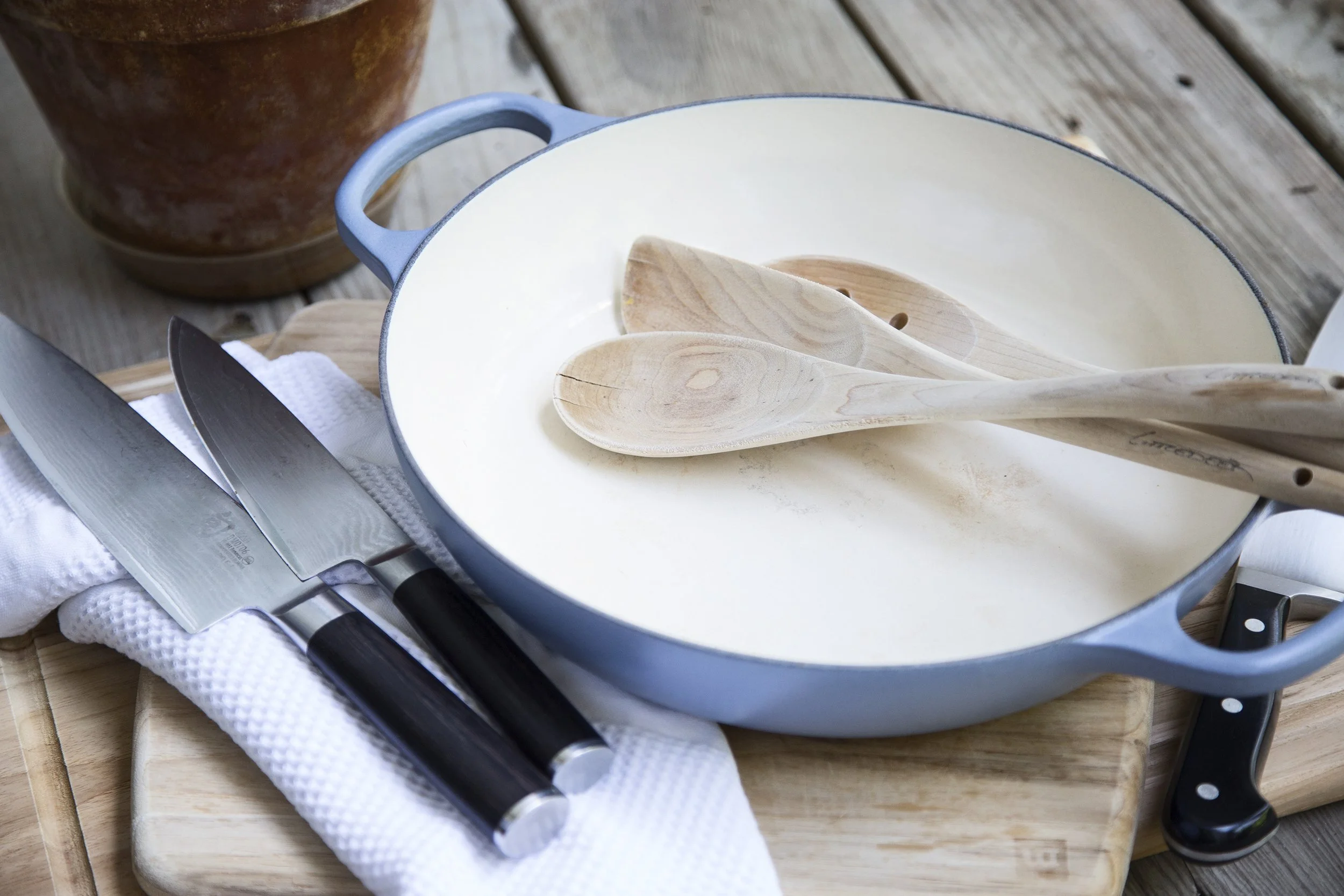 A blue enameled cast iron skillet with wooden spoons inside, placed on a wooden cutting board, with kitchen knives and a white cloth underneath, on a wooden table.