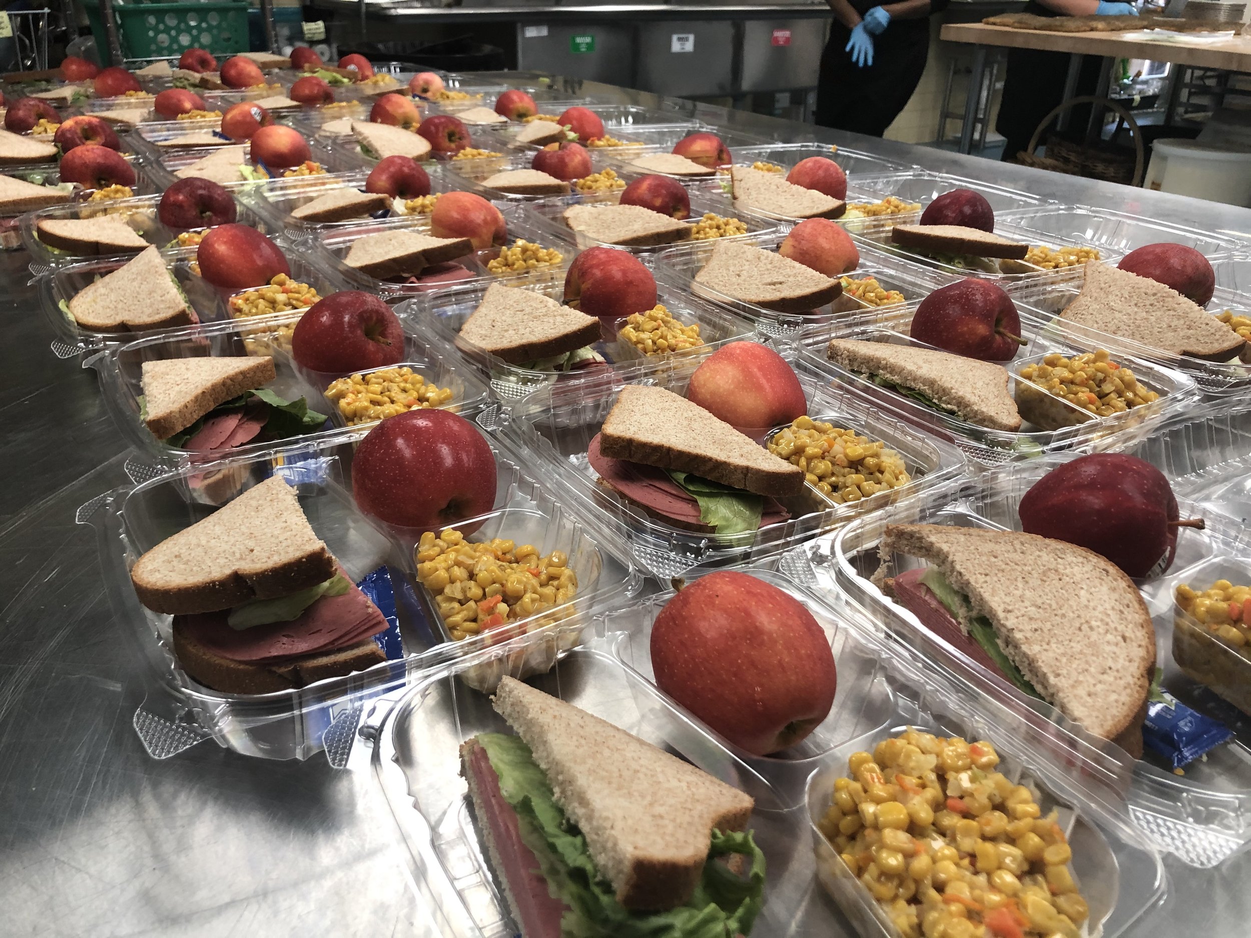 Multiple clear plastic meal trays arranged on a stainless steel table, each containing an apple, a sandwich with ham, lettuce, and whole wheat bread, and a side of corn salad with diced carrots and peppers.