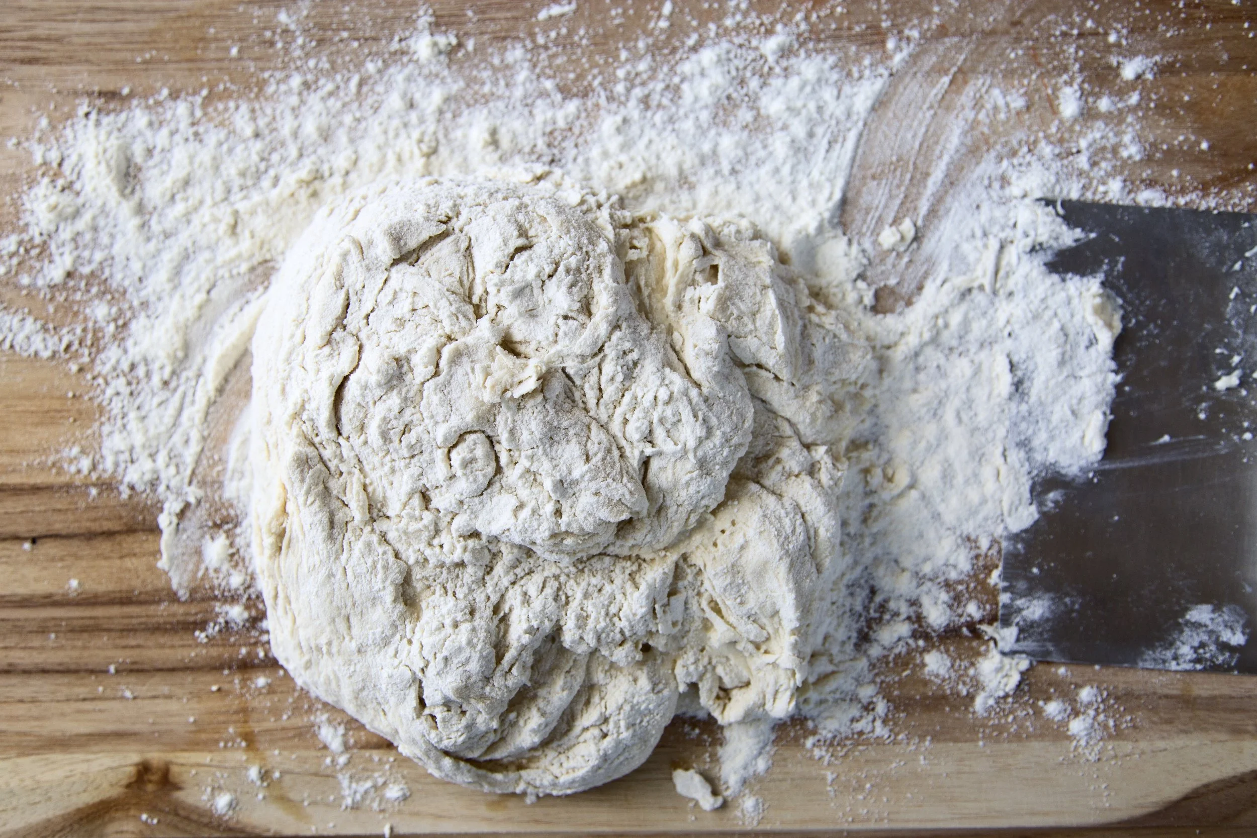Dough appears to be kneaded and is on a wooden surface with flour, a dough scraper, and a partially visible rolling pin.