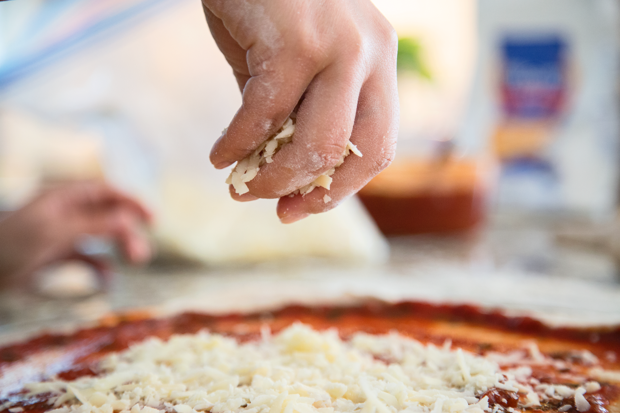 A hand sprinkling shredded cheese onto a pizza with tomato sauce. In the background, there are other cooking ingredients.
