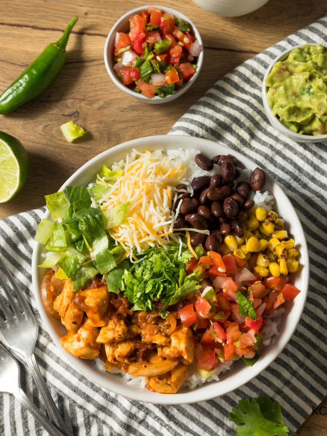 A bowl of rice topped with shredded cheese, grilled chicken in sauce, black beans, corn, chopped lettuce, cilantro, and a pico de gallo salsa. Additional bowls of guacamole and pico de gallo, lime halves, and a jalapeño pepper are on a wooden table with a striped napkin.