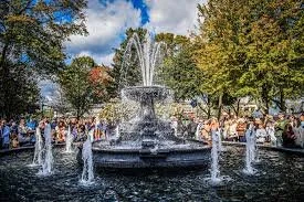Fountain in a park with people gathered around, trees with fall foliage, and a clear sky.