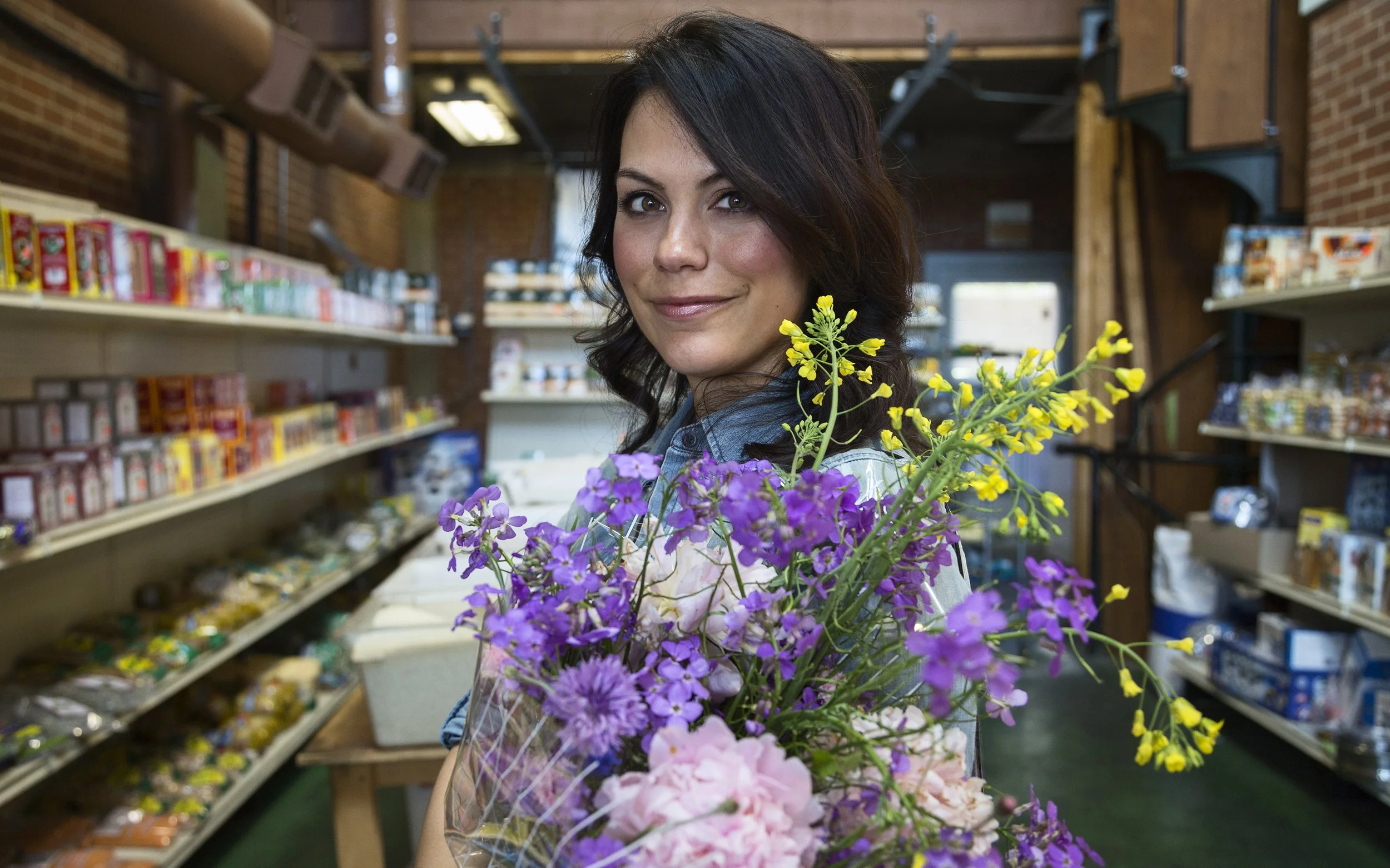 A woman with brown hair holding a bouquet of purple, pink, and yellow flowers inside a grocery store or flower shop.