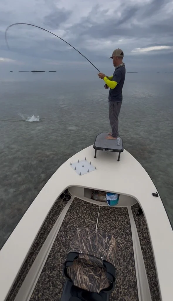 Fly angler standing on the bow of a technical poling skiff in the Florida Keys, hooked up to a powerful fish in shallow clear water. This flats fishing adventure highlights the thrill of sight fishing for tarpon, snook, and redfish in pristine shallo