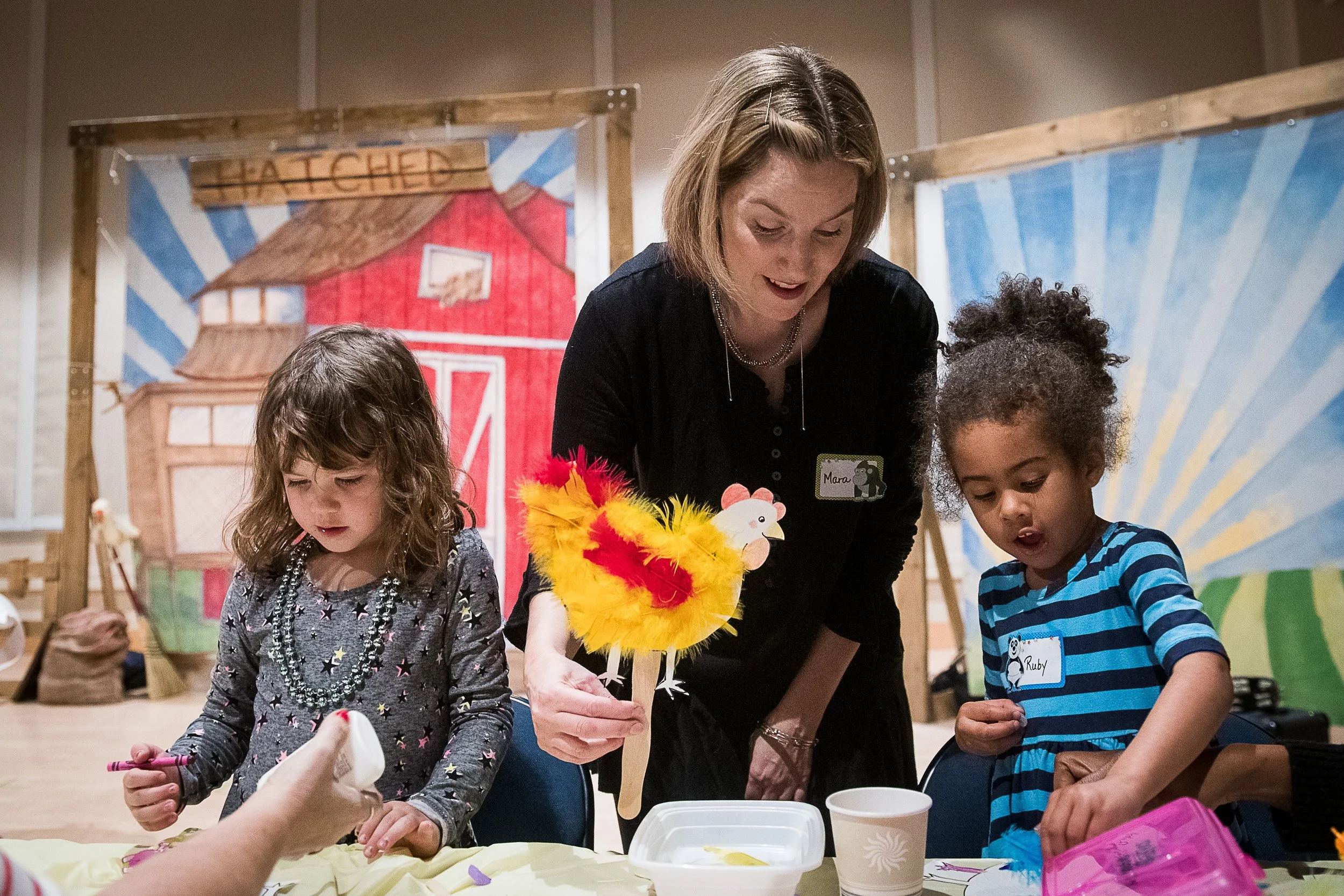 A woman with short blonde hair helping two young girls with craft activities at a table, with a colorful barn-themed backdrop behind them.