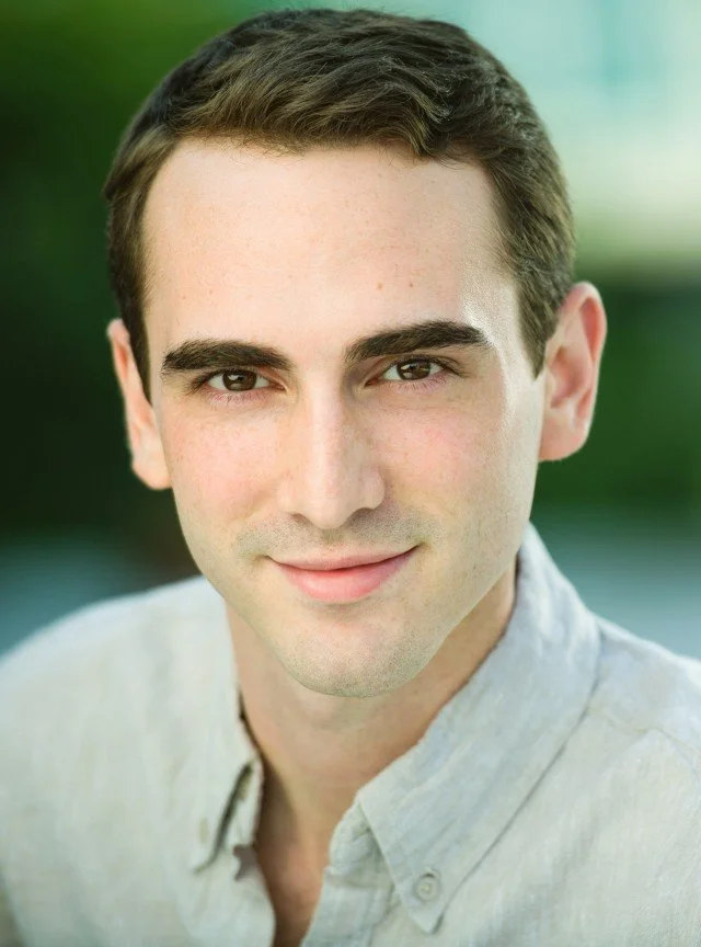 A young man with short dark hair and light skin, smiling, wearing a light-colored button-up shirt, outdoors with a blurred green background.