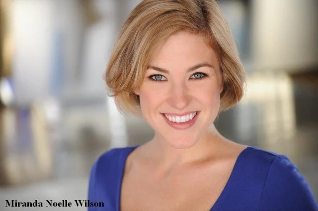A smiling woman with short blonde hair and blue eyes wearing a blue top, standing indoors with a blurred background.