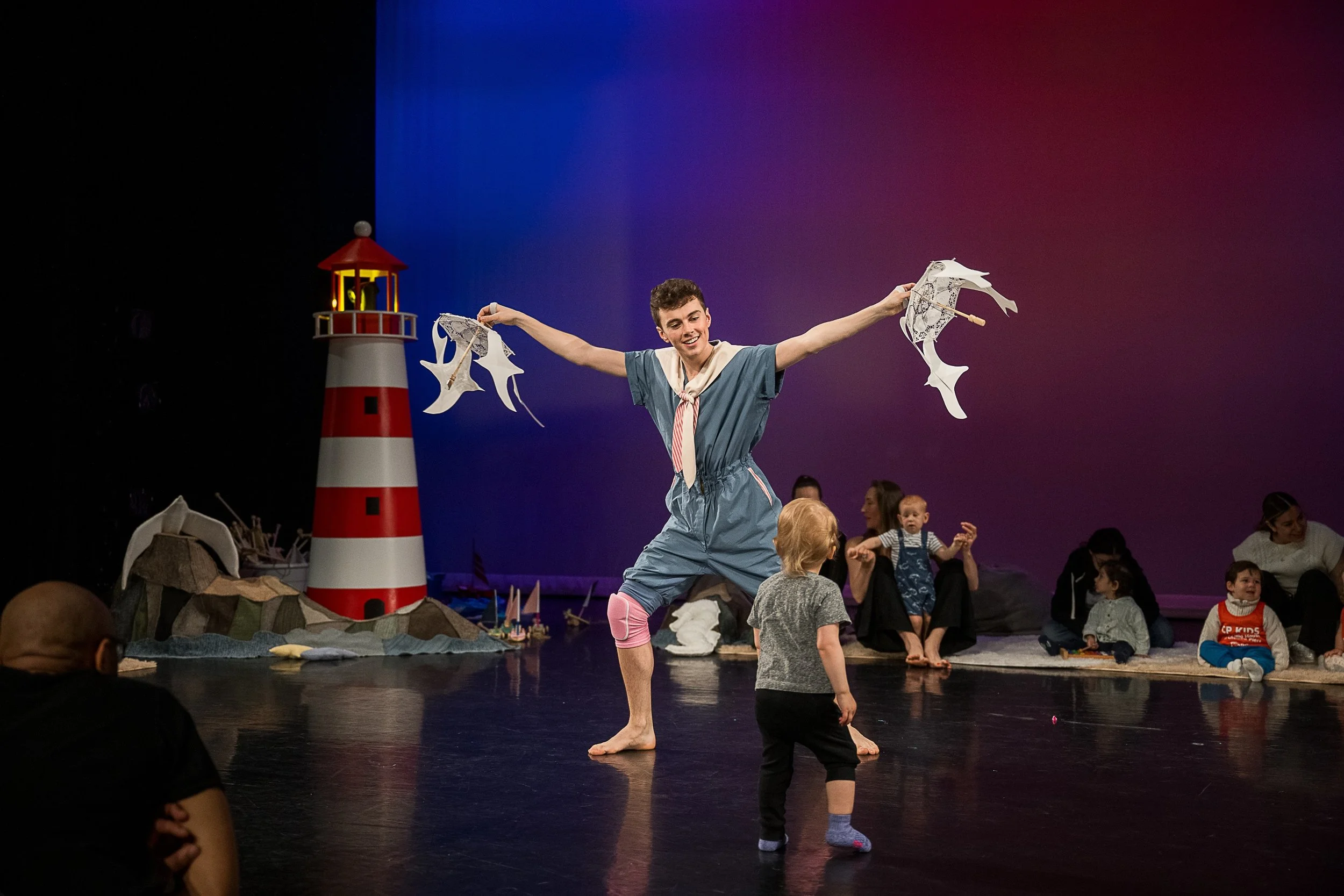 Performer in a blue costume with a white necktie dancing on stage with children watching, a lighthouse prop and small sailboats in the background