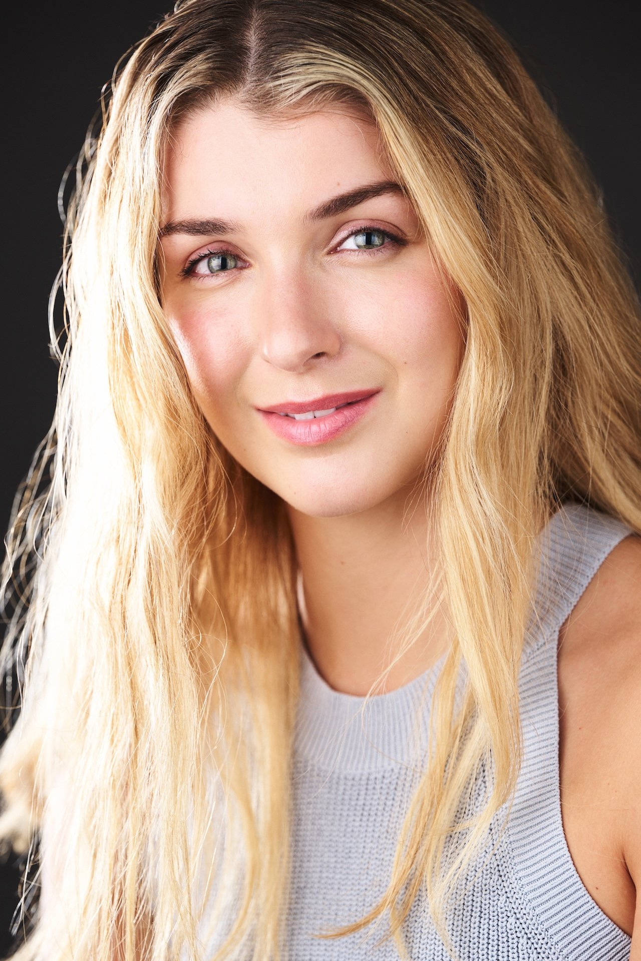 A young woman with long blonde hair, blue eyes, and a light gray top, smiling softly at the camera.