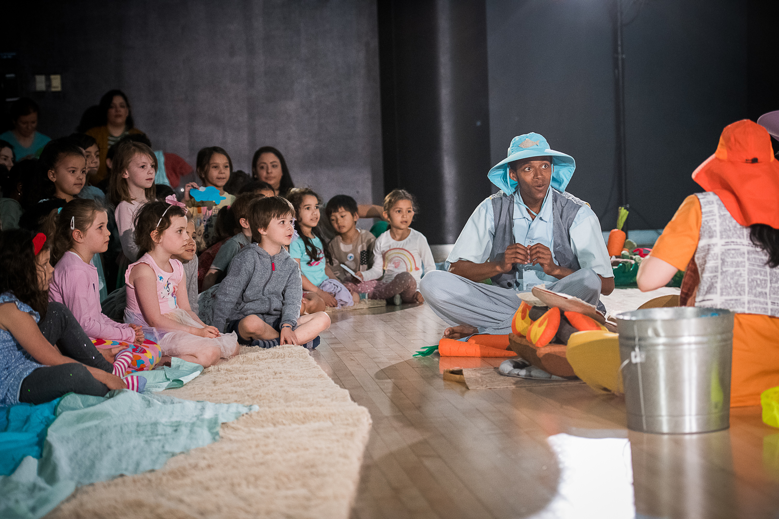 A man dressed as a farmer in a blue hat and overalls entertains a group of children sitting on the floor during a puppet show about a farm scene with carrots and other vegetables.