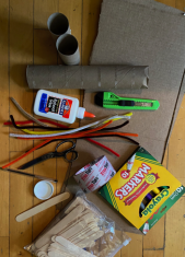 A small workspace with electronic tools and supplies: a pair of pliers, a bottle of super glue, a green utility knife, a bundle of multicolored wires, a white plastic component, and a yellow box of colored pencils on a wooden surface.