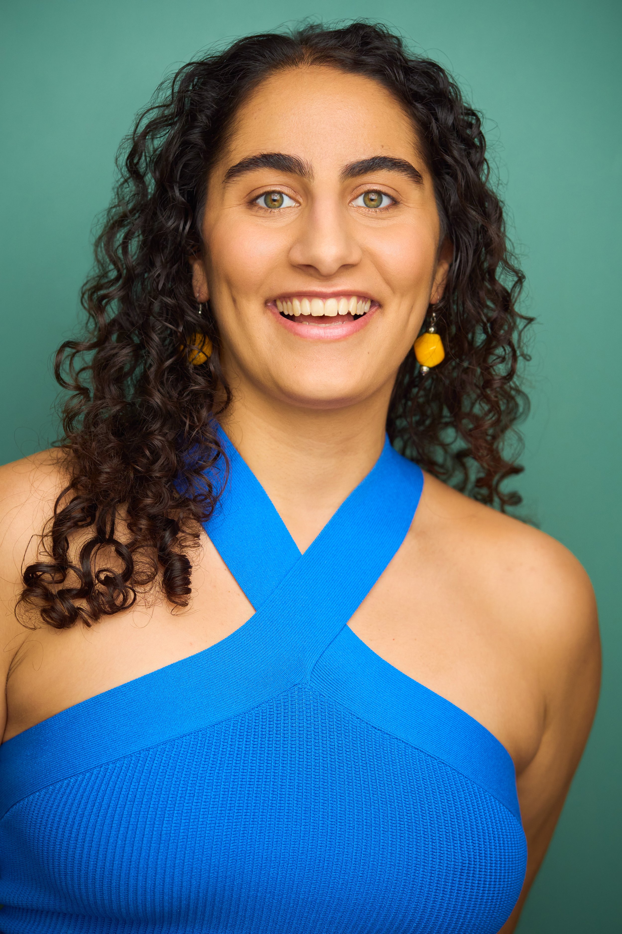 Portrait of a woman with long, curly red hair and blue eyes, wearing a dark green top, against a light-colored background.