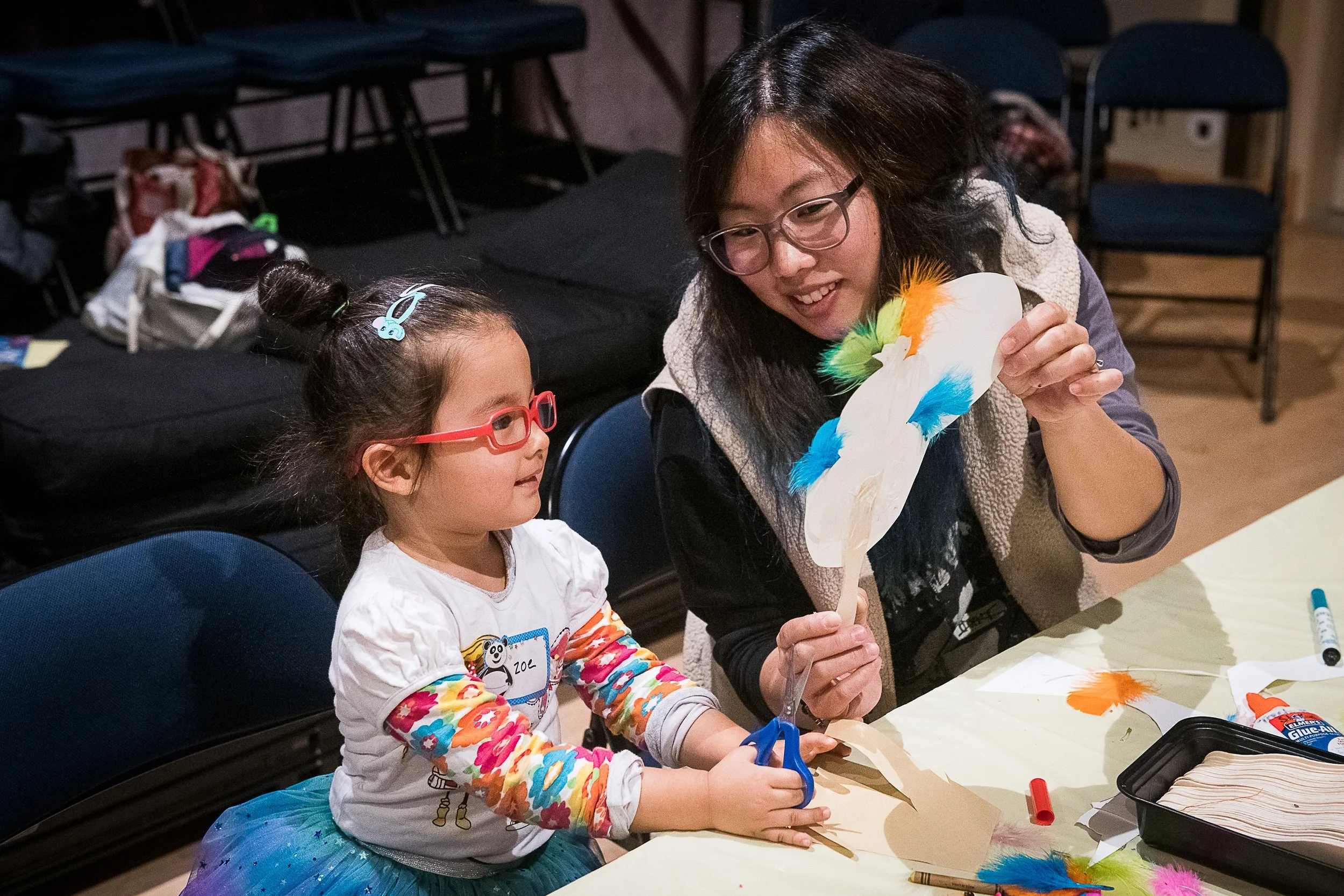 A woman and young girl sit at a table, making a colorful paper craft. The woman holds a decorated paper object with feathers, and the girl, wearing glasses and a colorful long-sleeve shirt, looks on. Art supplies and feathers are on the table.