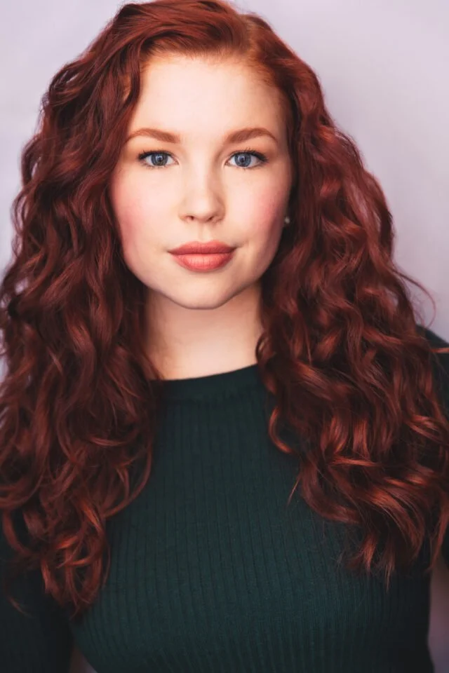 Portrait of a woman with long, curly red hair and blue eyes, wearing a dark green top, against a light-colored background.