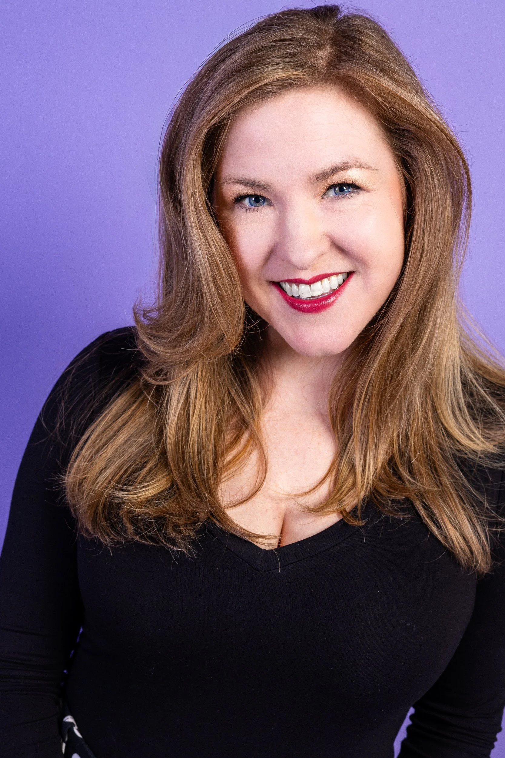 A smiling woman with long blonde hair and blue eyes wearing a blue top, standing indoors with a blurred background.