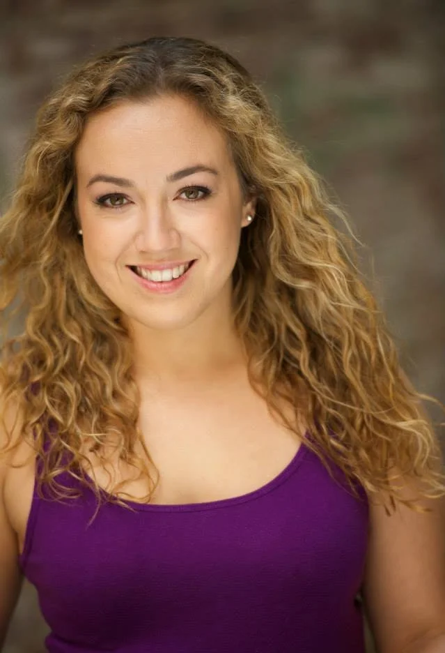 A woman with curly blonde hair smiling, wearing a purple top.