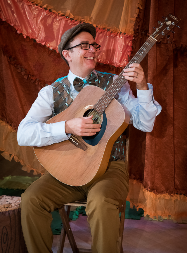 Man wearing glasses, a gray cap, and a bow tie, playing an acoustic guitar, smiling, in front of a decorated brown and orange curtain.