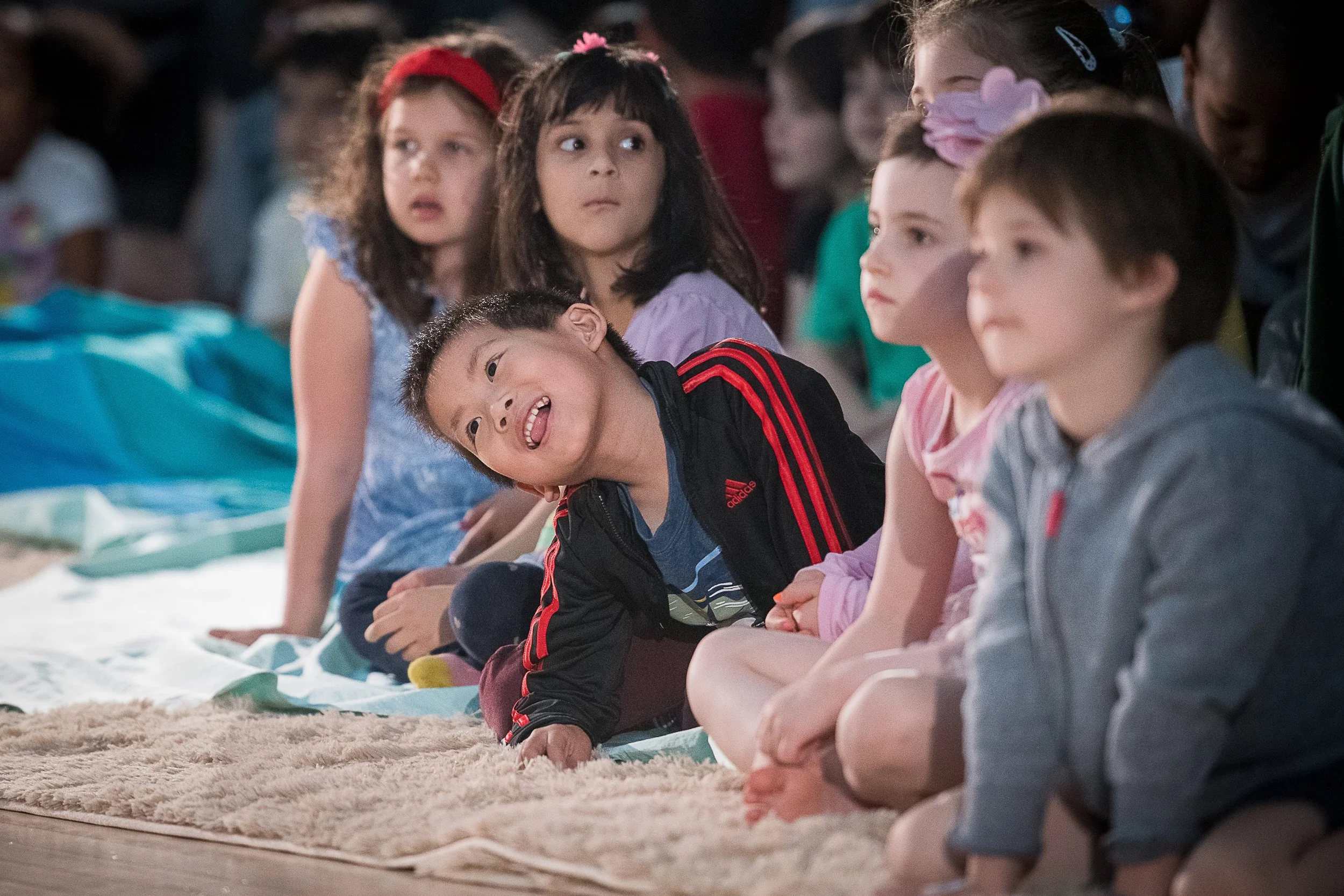 Children sitting on a carpet, some looking surprised or curious, during an outdoor event at night.