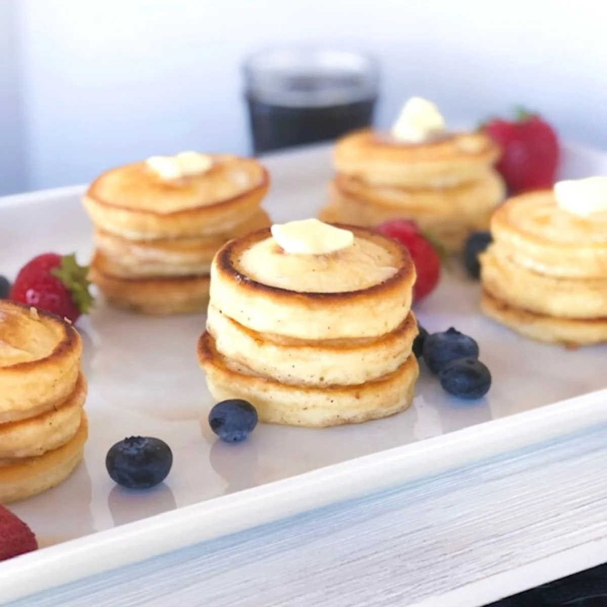Stacked mini pancakes topped with butter and served with fresh strawberries and blueberries on a white tray, with a glass of dark beverage in the background.