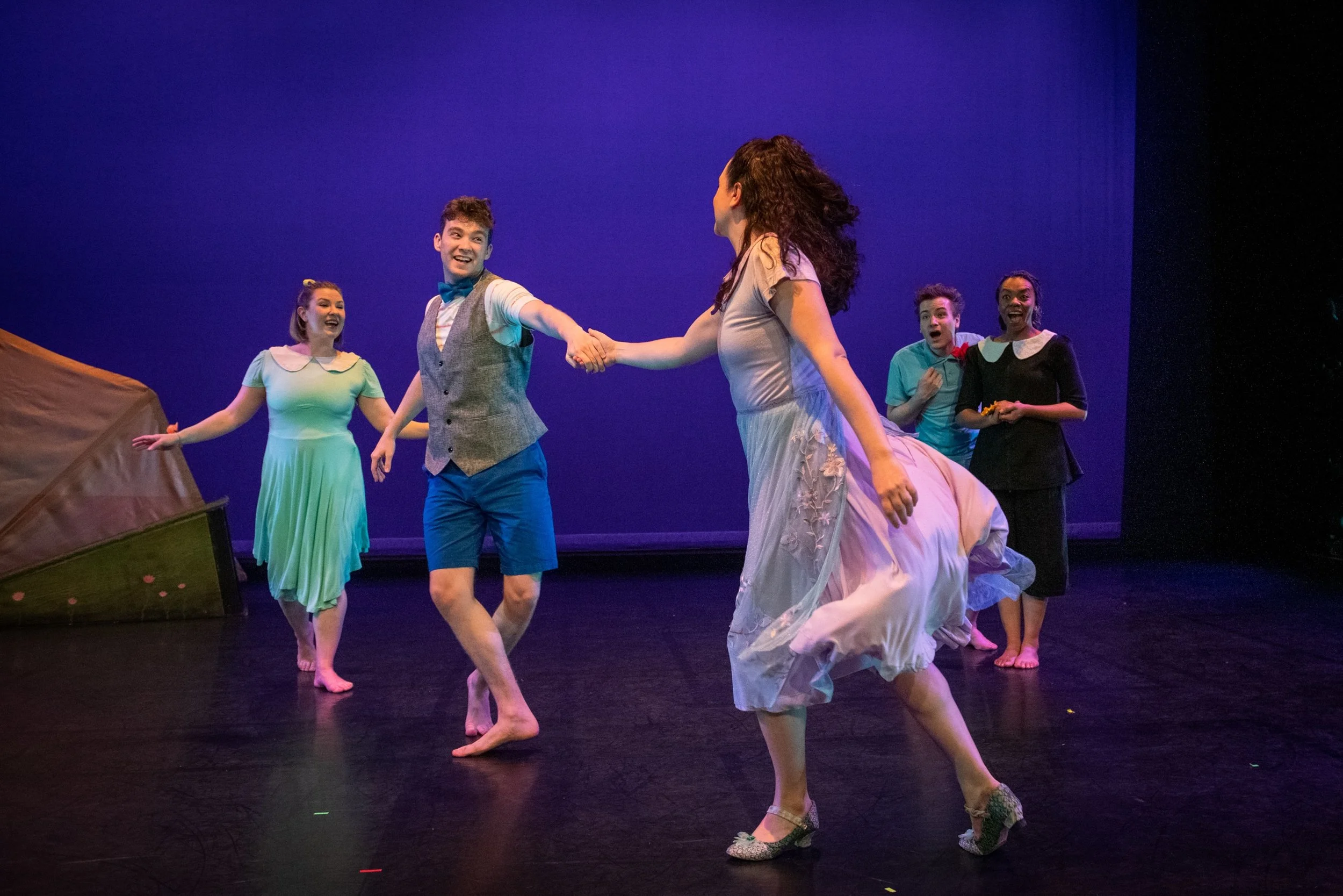 Actors performing on stage in a theatrical production, with one actor holding hands with another while four others observe and smile, set against a plain purple background.