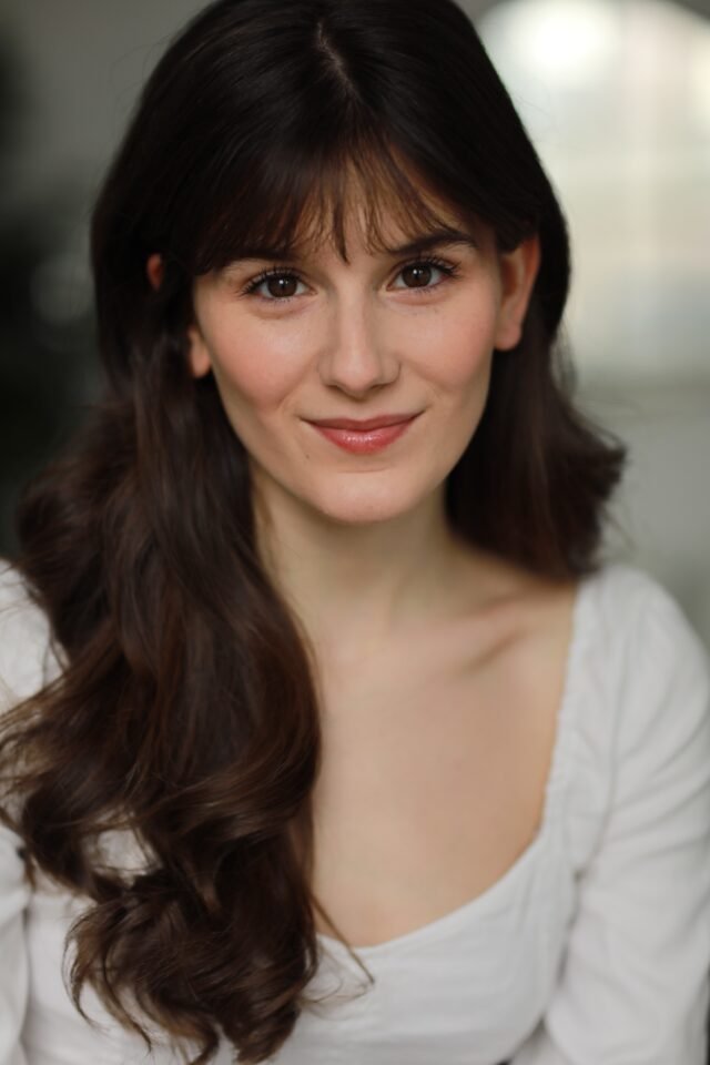 A young woman with long, dark brown hair and a friendly smile, wearing a white top, standing outdoors with a blurred background.