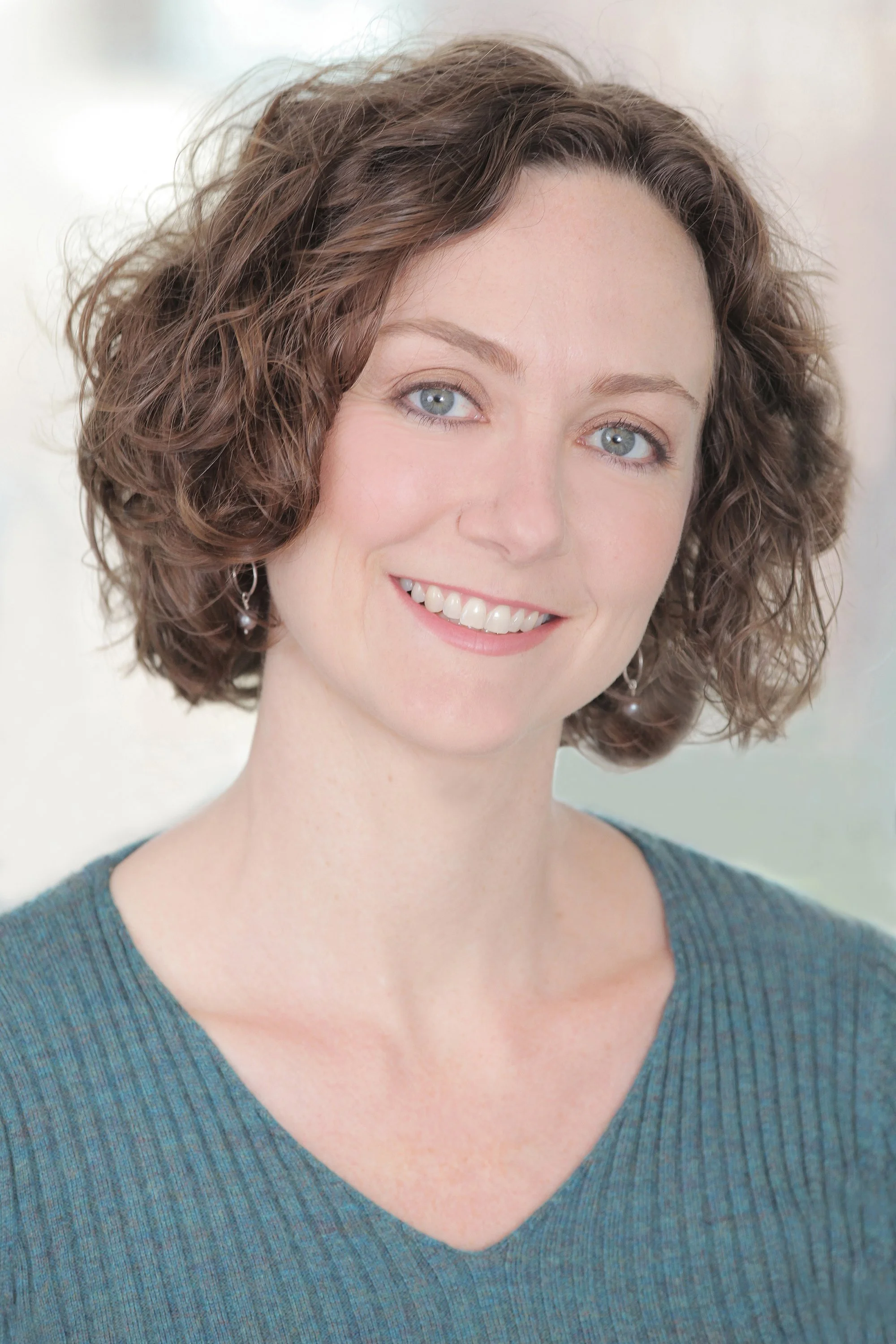 Headshot of a woman with short, curly brown hair, blue eyes, wearing a blue V-neck top and earrings, smiling at the camera.