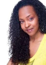 A woman with curly black hair and a yellow top, smiling softly at the camera.