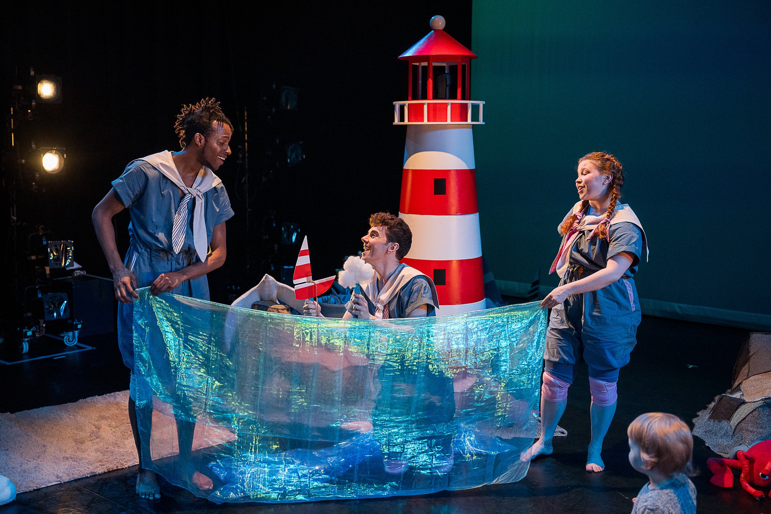 Three children in sailor costumes performing a play on stage with a lighthouse prop, a blue ocean-like sheet, and ocean-themed accessories, with a young child watching in the foreground.