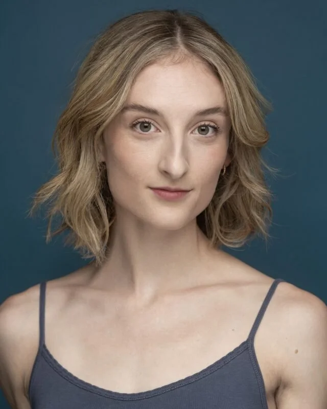 A young woman with shoulder-length blonde hair and light skin, wearing a dark gray spaghetti strap top, looking at the camera with a slight smile against a solid dark blue background.