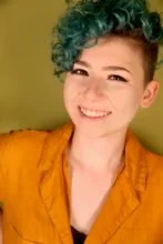 Young woman with short dyed green curly hair smiling, wearing an orange jacket against a green background.